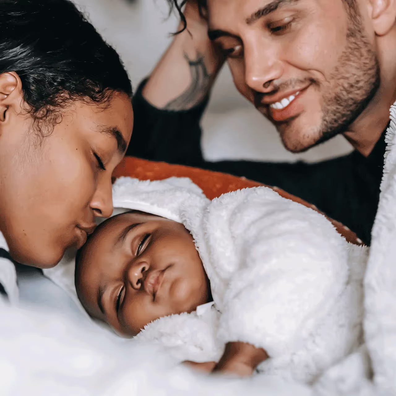 A mother and father affectionately looking at their sleeping baby, who is wrapped in a blanket