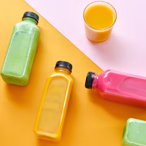 Colorful detox juice bottles, including green, yellow, and pink drinks, arranged on a bright yellow and pink background.