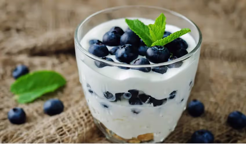 A glass of yogurt layered with fresh blueberries, topped with a mint leaf on a rustic background.