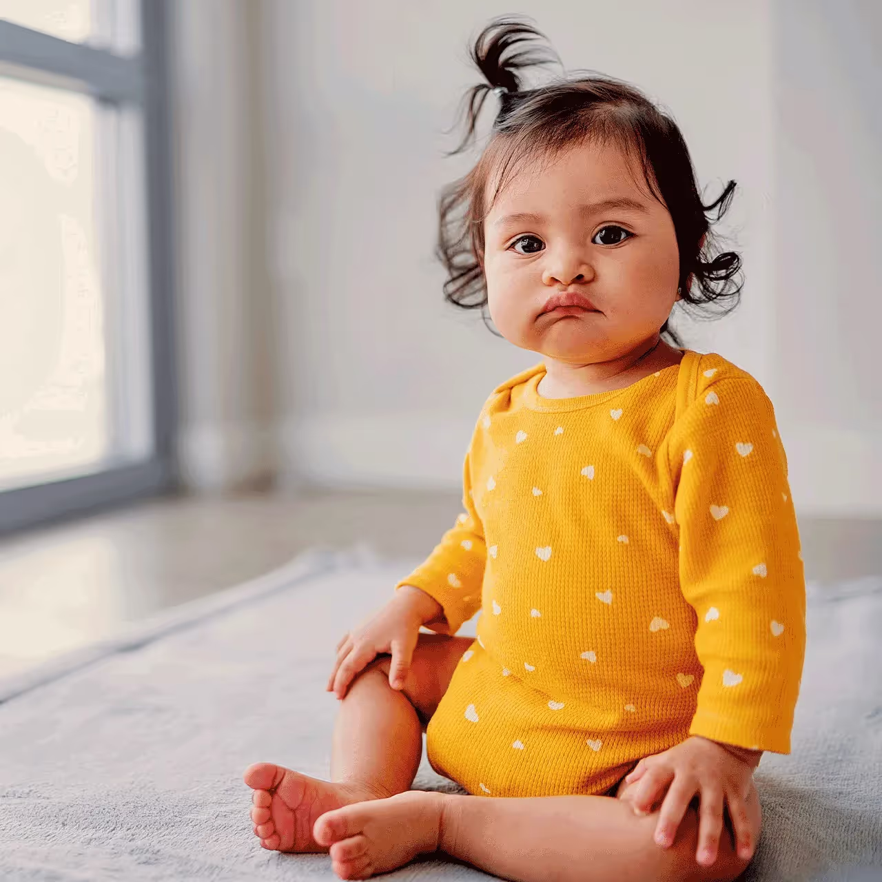 Baby sitting on the floor, wearing a yellow onesie with white hearts, looking curiously at the camera