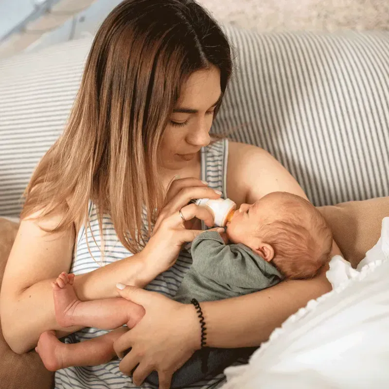 A mother feeding her baby a bottle containing human milk oligosaccharides while sitting on a couch
