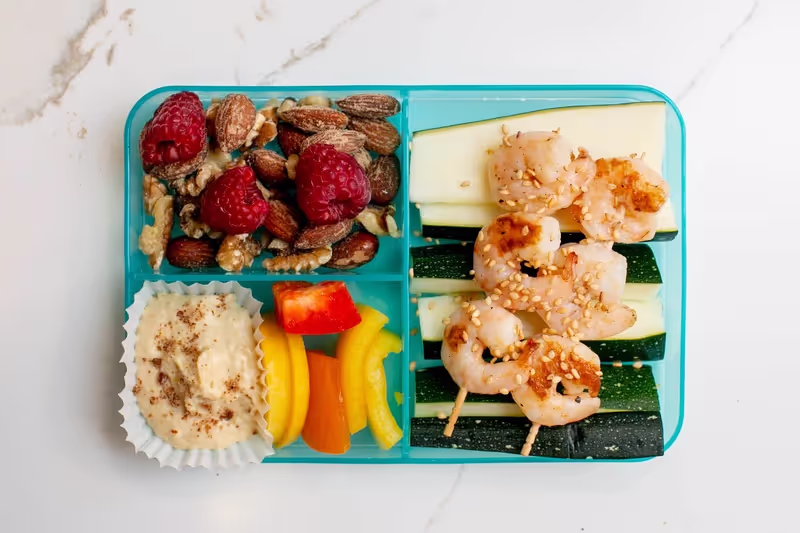 A rainbow-inspired bento box lunch with nuts, raspberries, zuchinni, sesame shrimp skewers, bell peppers and hummus sprinkled with flax