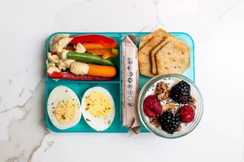 A rainbow-inspired bento box lunch with yogurt, berries, granola, chia, crackers, veggies, hummus, a hard-boiled egg, and a packaged granola bar