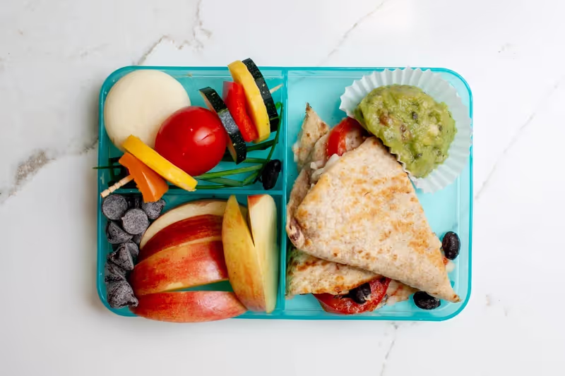 A rainbow-inspired bento box lunch with tomato and black bean cheese quesadillas, guacamole, apple, cheese, a veggie skewer, and dark chocolate chips