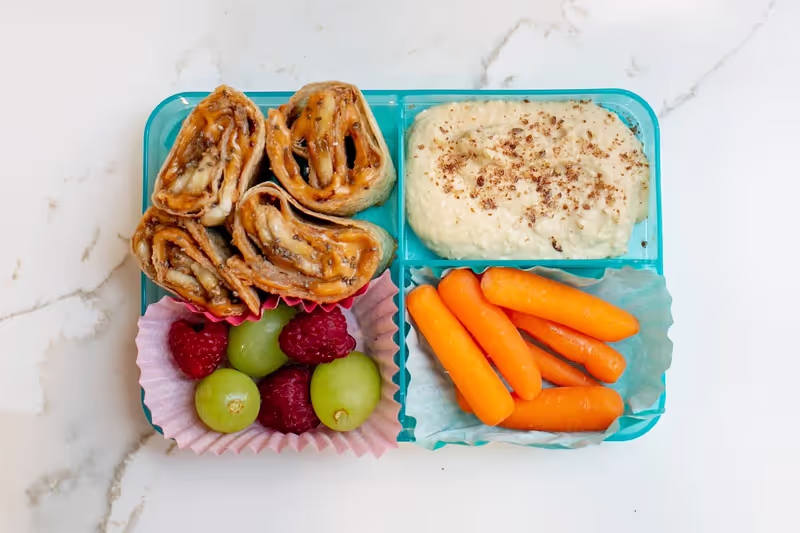 A rainbow-inspired bento box lunch with nut/seed butter banana wraps, raspberries, grapes, carrots and hummus sprinkled with flax seed