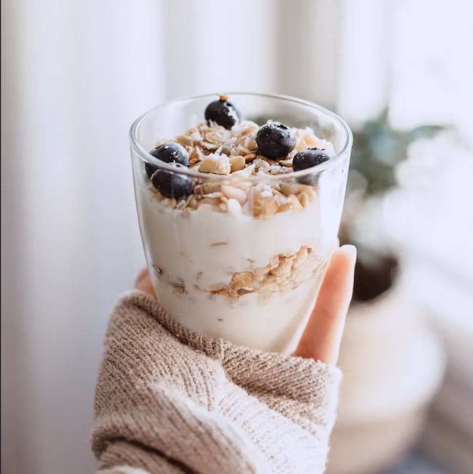 Close-up of a hand holding a glass of yogurt with granola and blueberries, pondering if it is suitable for babies