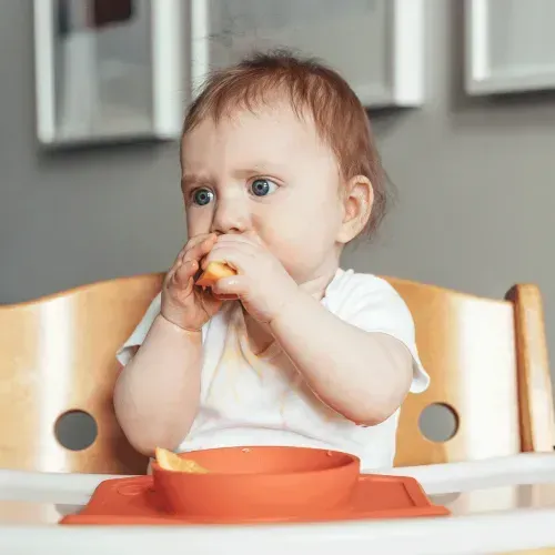 Baby in high chair eating a fruit, representing when to introduce common allergens, like peanut butter, to babies