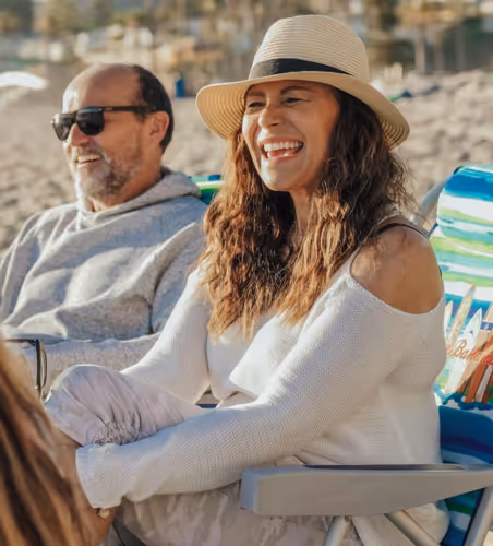 A midlife woman and man relax on beach chairs in the sand, smiling and enjoying a sunny day by the ocean.
