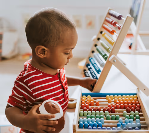 Toddler playing with colorful abacus at daycare, wearing a red striped shirt and holding a toy football in a bright classroom.