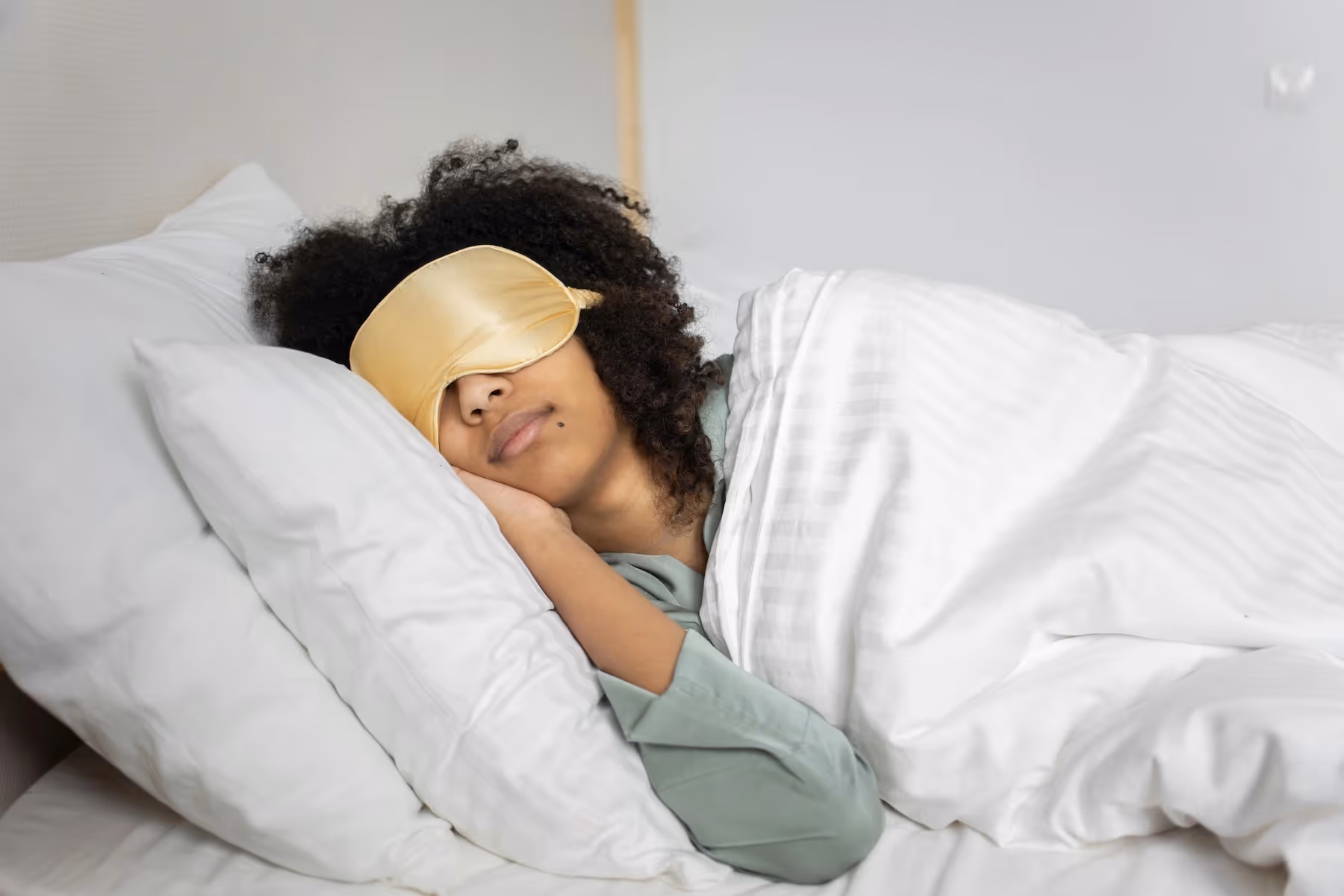A woman of color sleeping in bed wearing a gold sleep mask, wrapped in a white blanket.