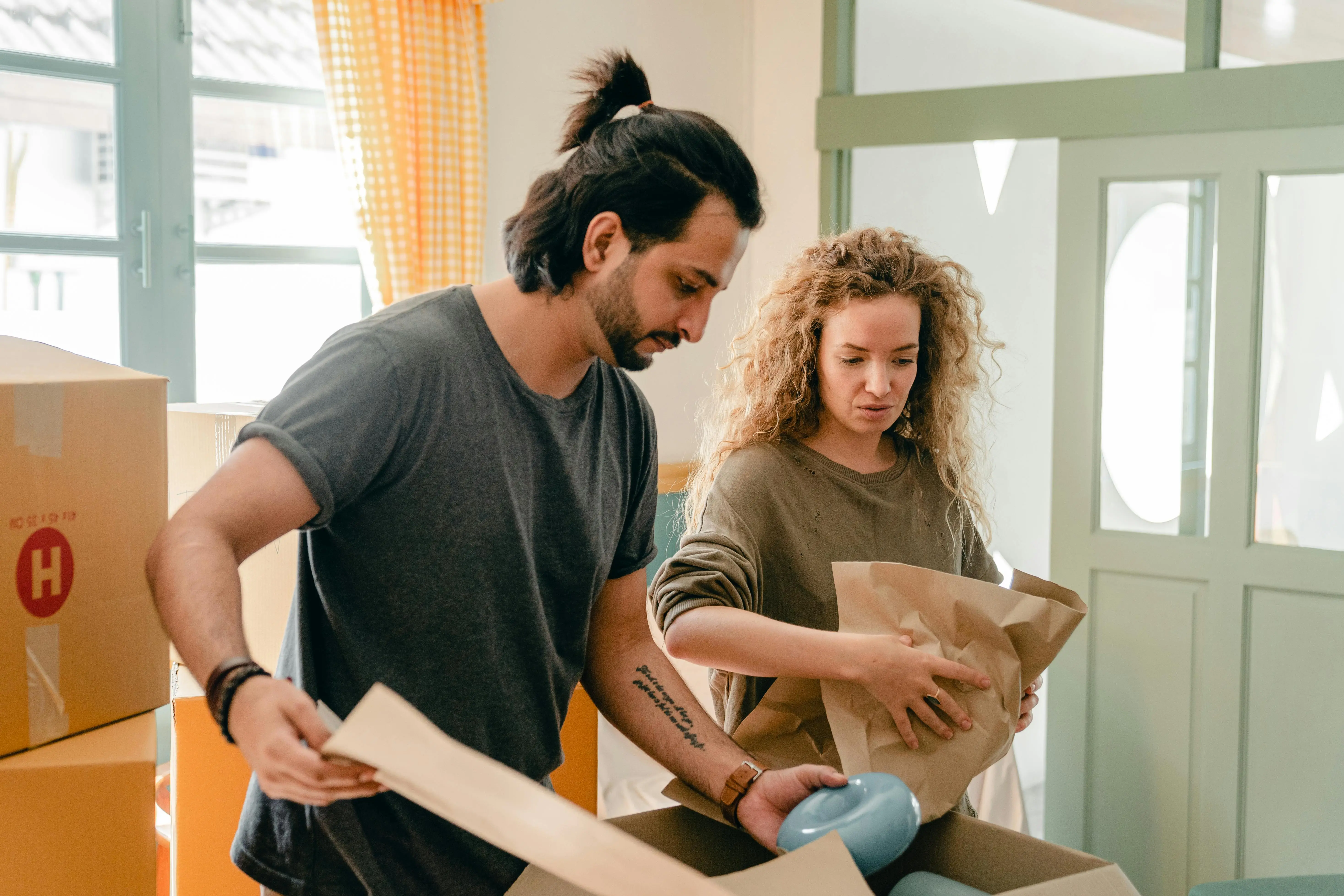 A couple unpacking or packing household items in a well-lit room with moving boxes.