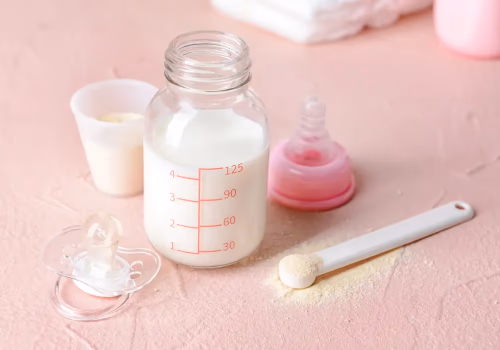 A baby bottle with infant formula, spoon with powder formula, and pacifier on a table.