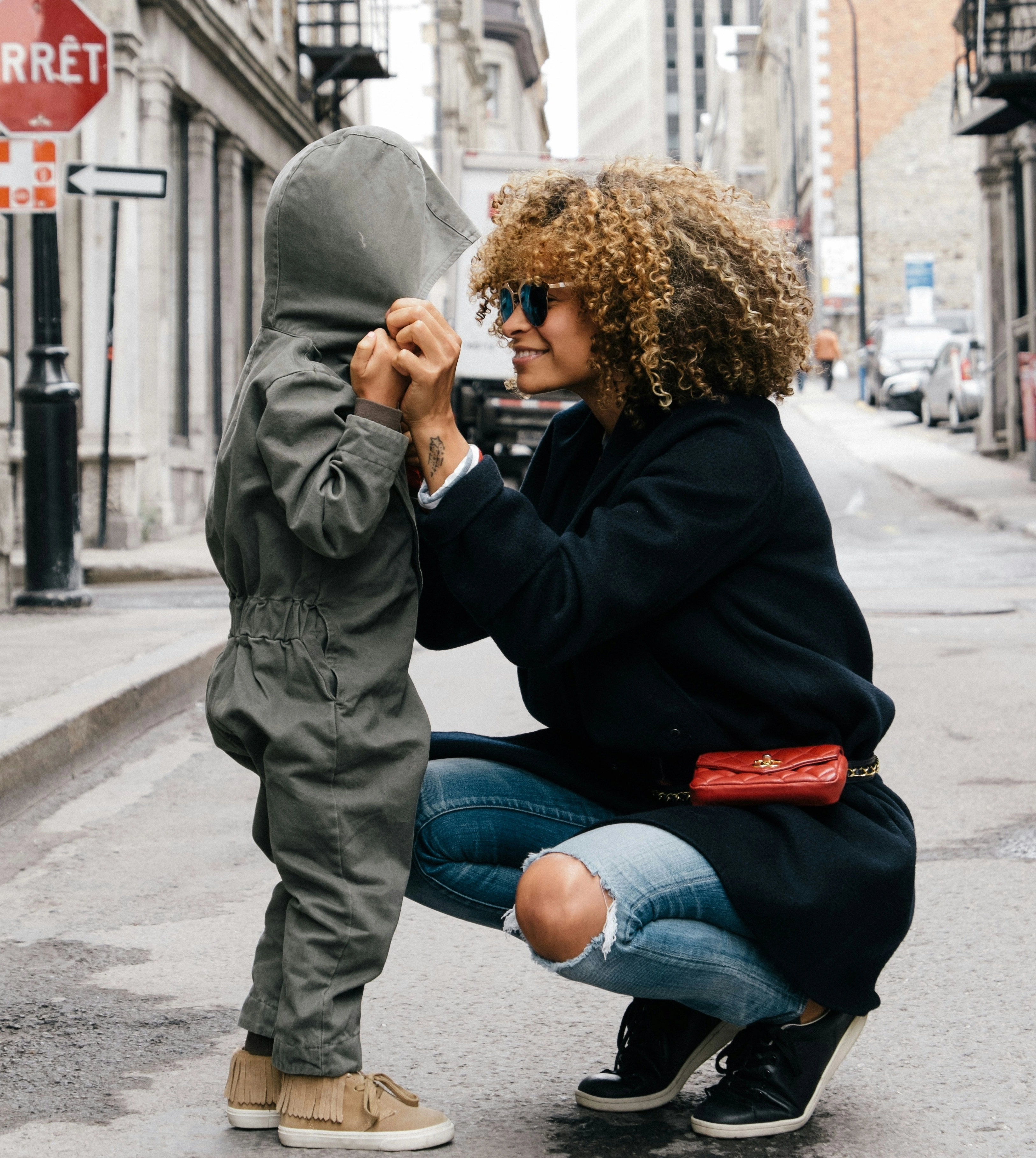  mother adjusting her child’s hood on a foreign city street, smiling while wearing sunglasses and a dark 