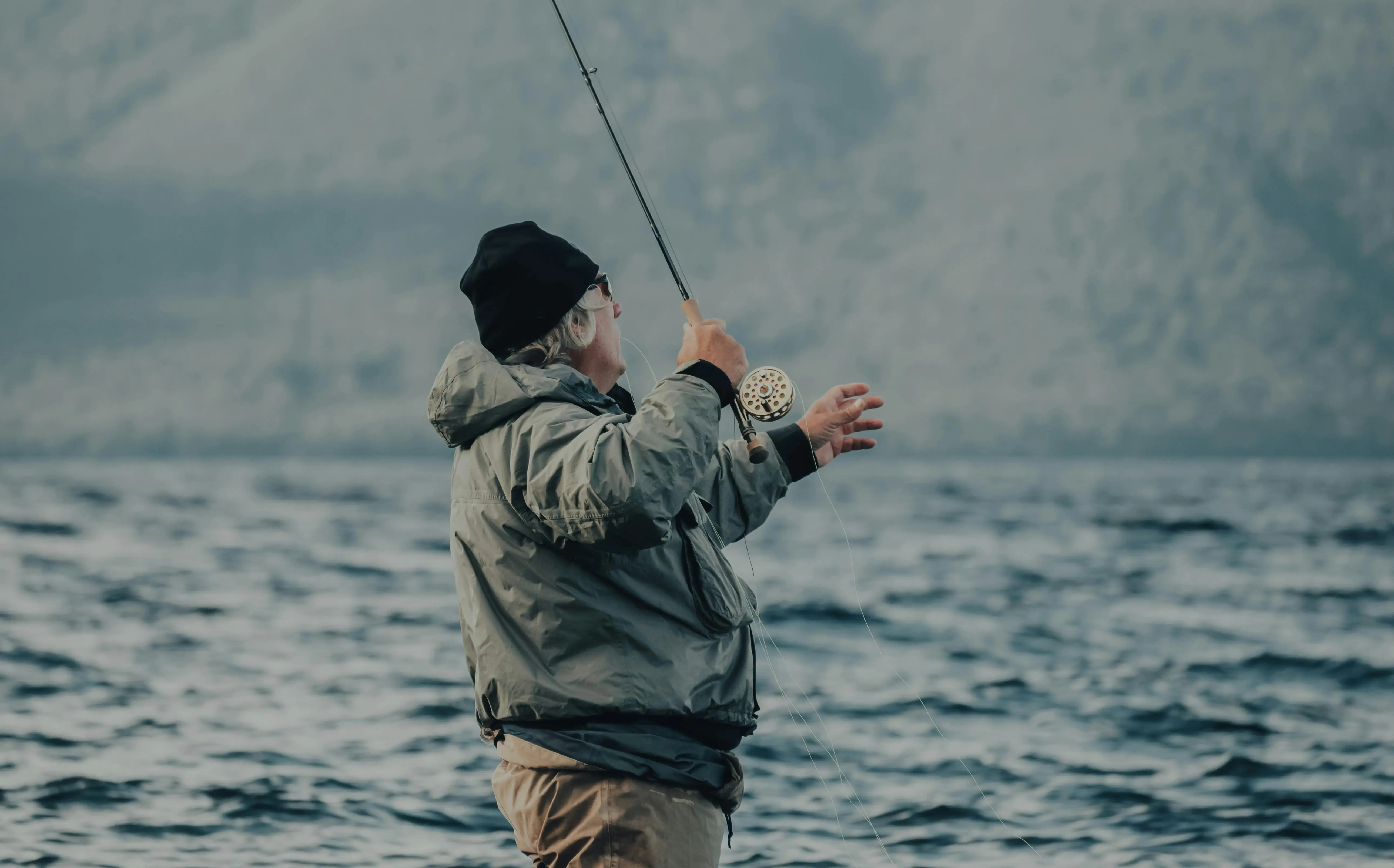 A gray-haired man fly-fishes alone on a lake, surrounded by mountains and water.