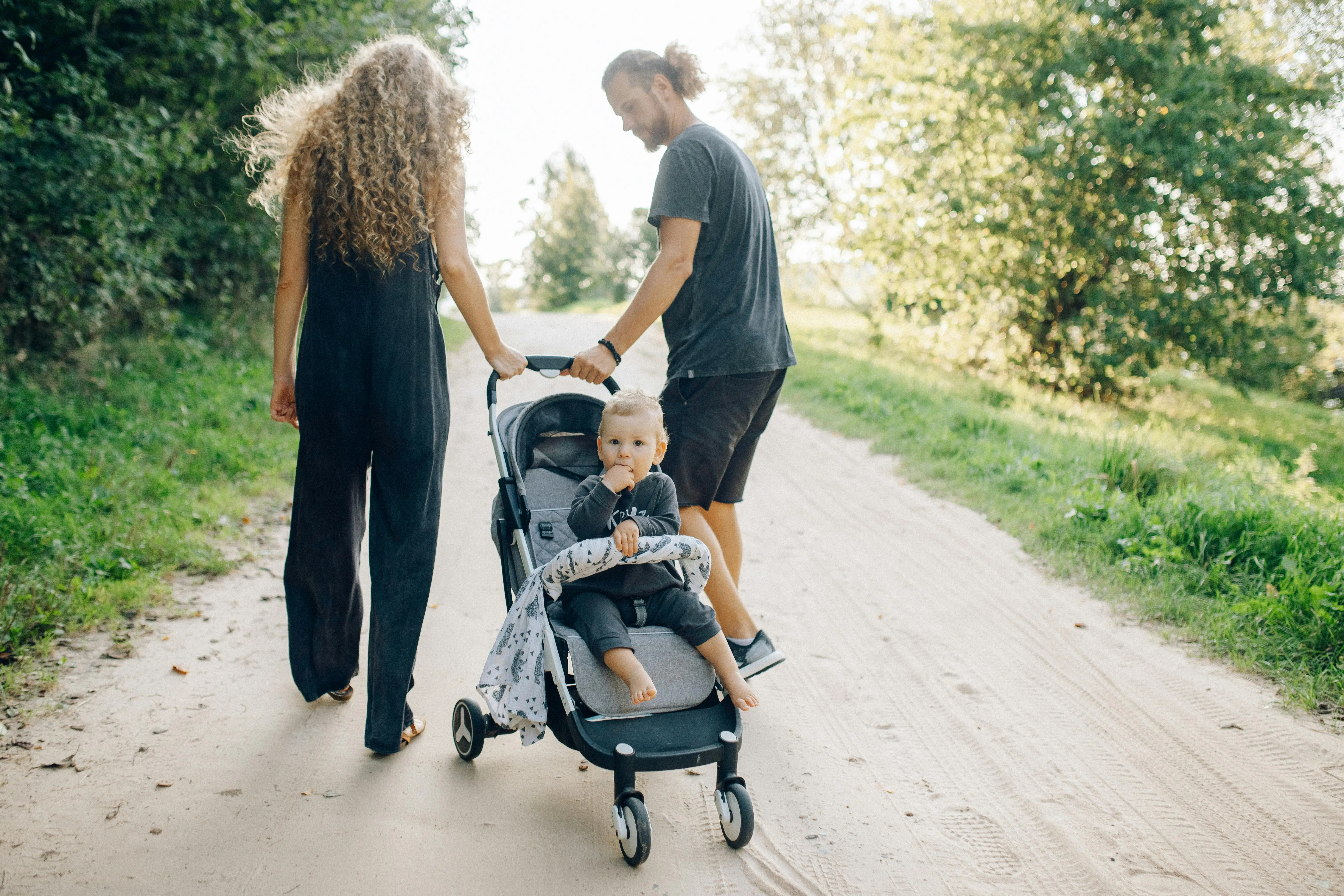 A couple walks outdoors pushing a stroller with a toddler, surrounded by greenery.