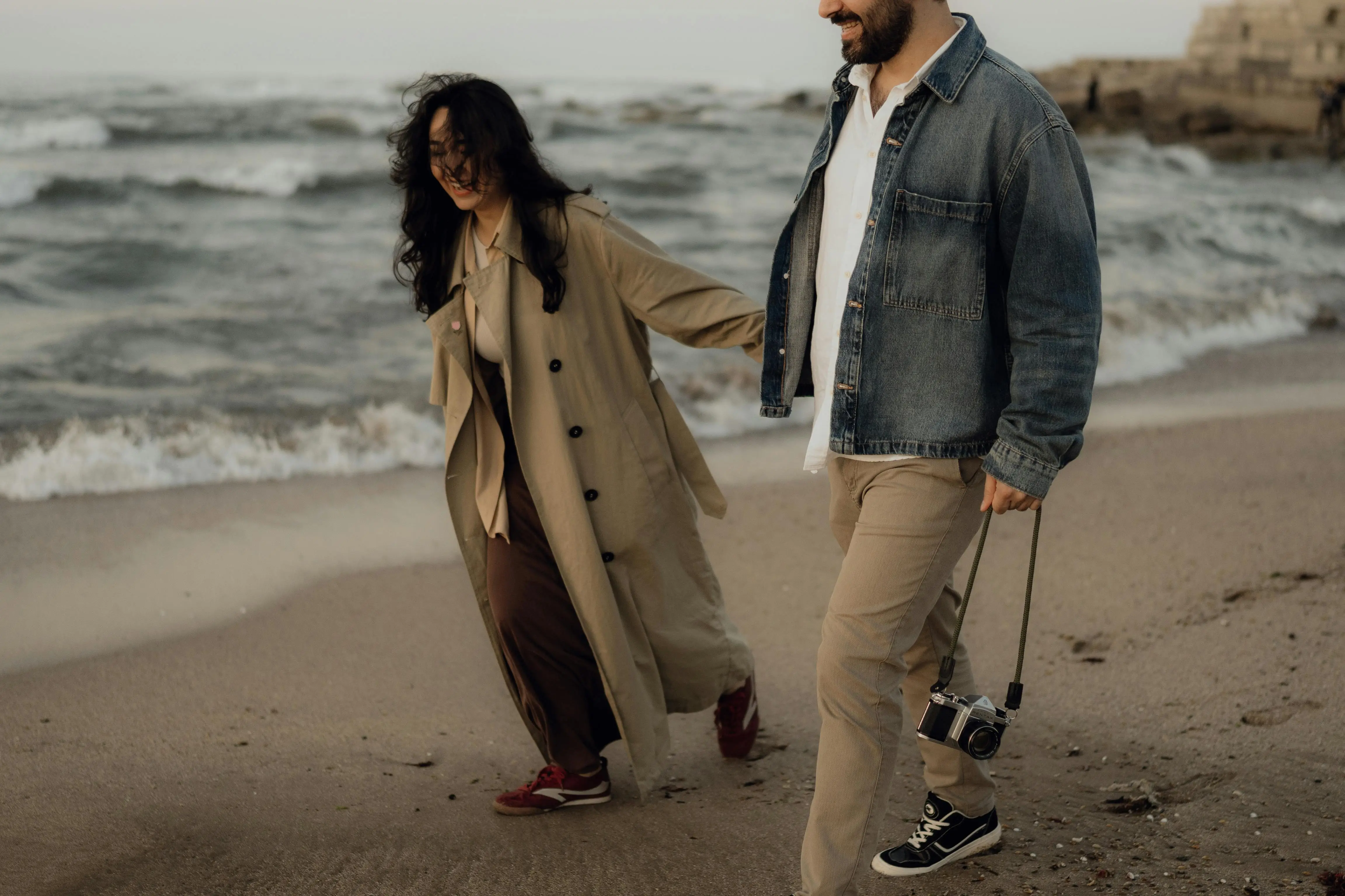 A smiling couple walks hand in hand along the beach, one holding a camera.