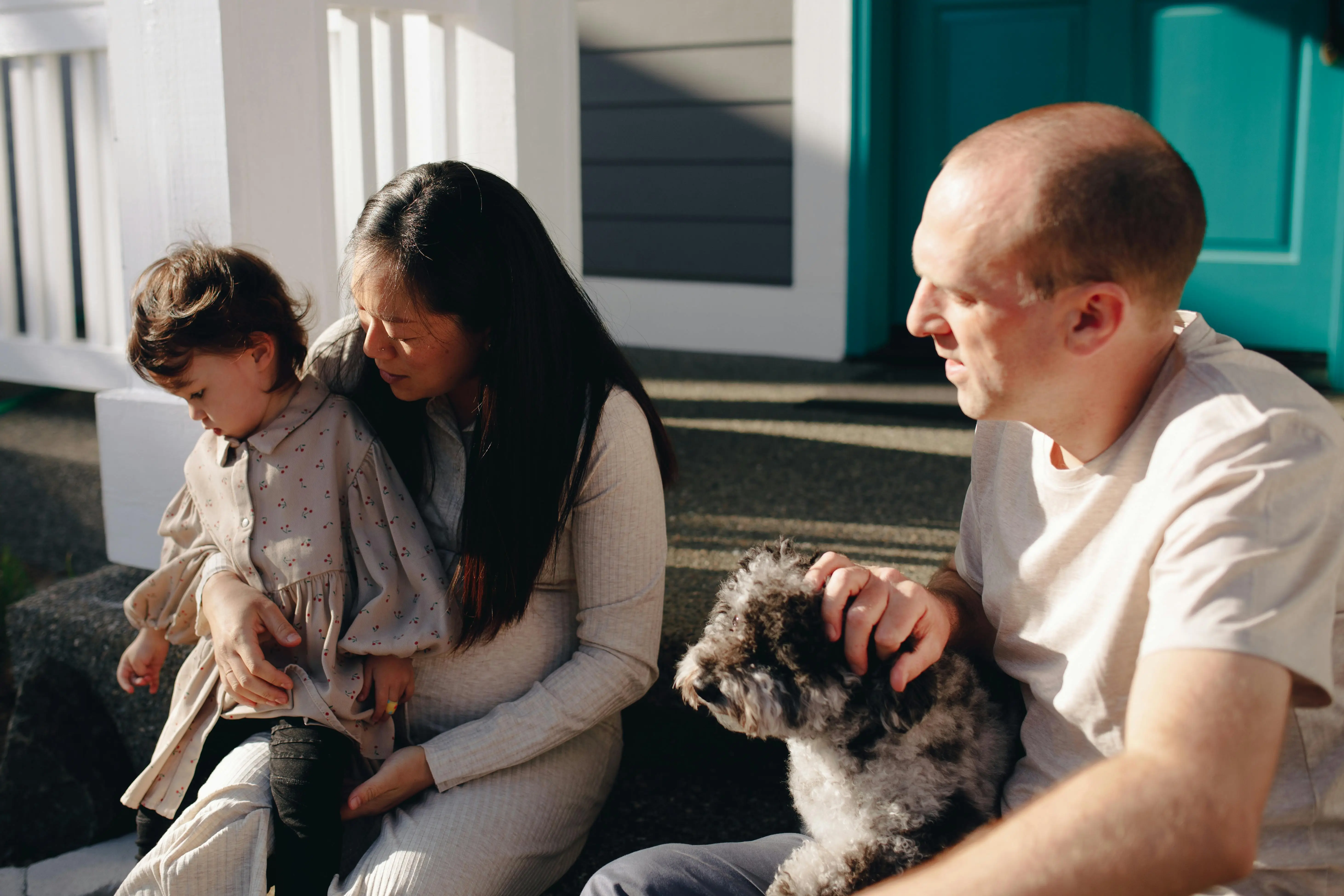 A couple sits on a porch with their child and dog, relaxing in the sunlight.
