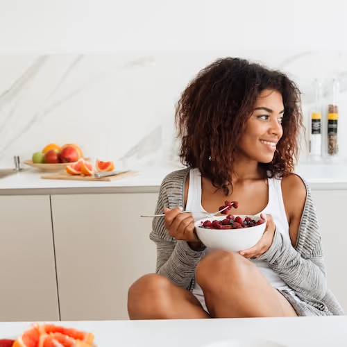 Smiling woman sitting at kitchen counter holding bowl of fruit, in bright white kitchen.