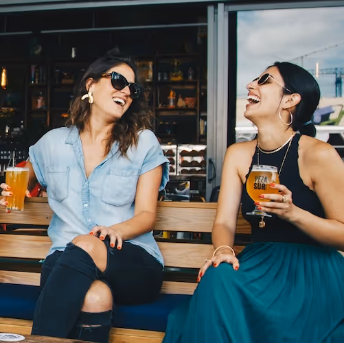 Two young women sit on an outdoor bench, laughing and holding their glasses of beer.