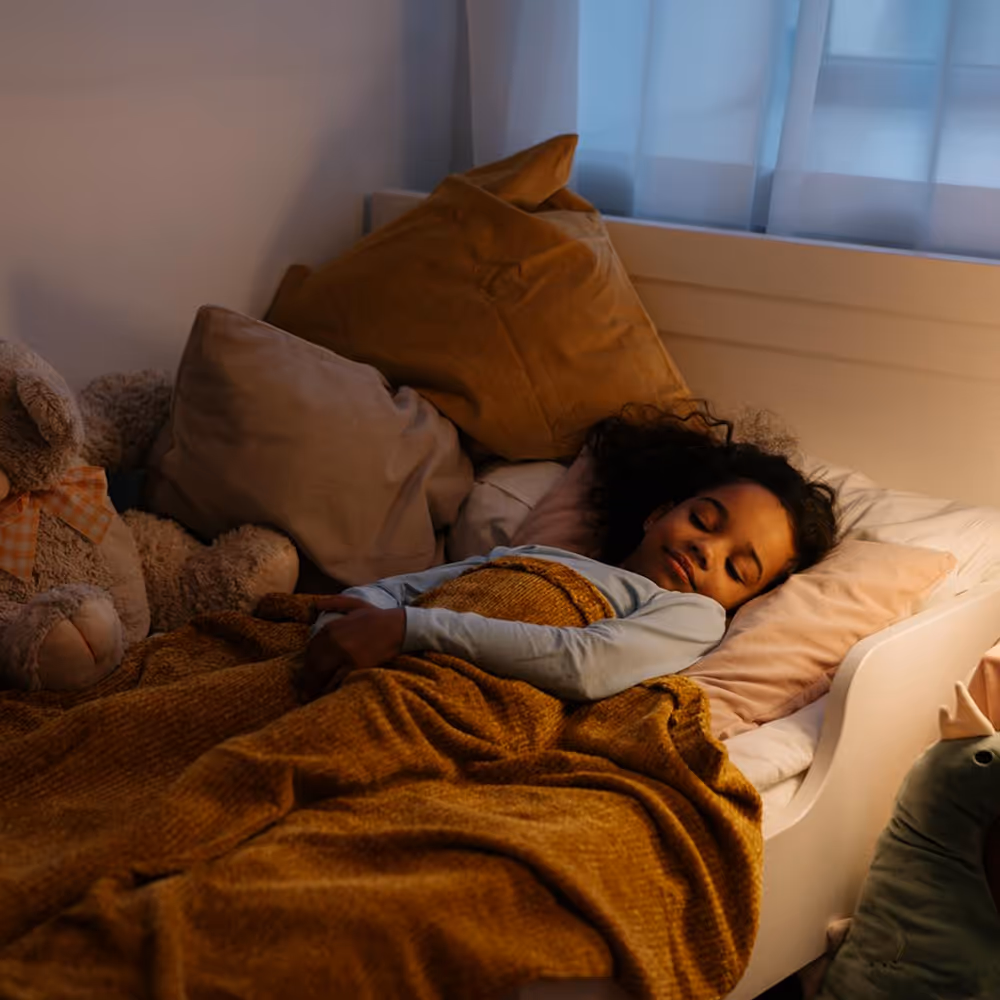 Young girl peacefully sleeping in bed with a mustard yellow blanket, surrounded by pillows and stuffed animals.