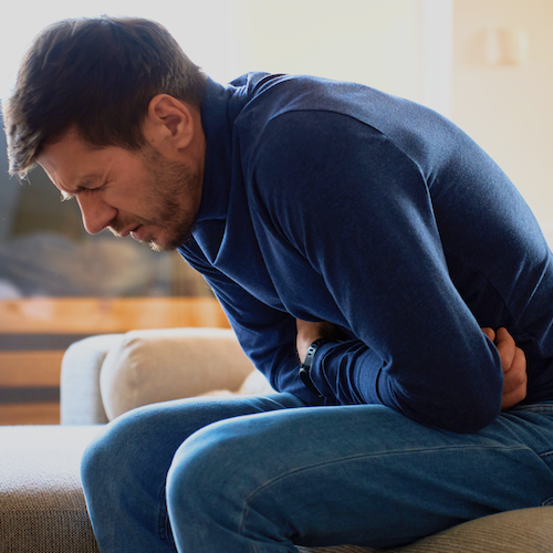Man sitting on couch hunched over in pain, holding his stomach with both hands, wearing a blue long-sleeve shirt and jeans