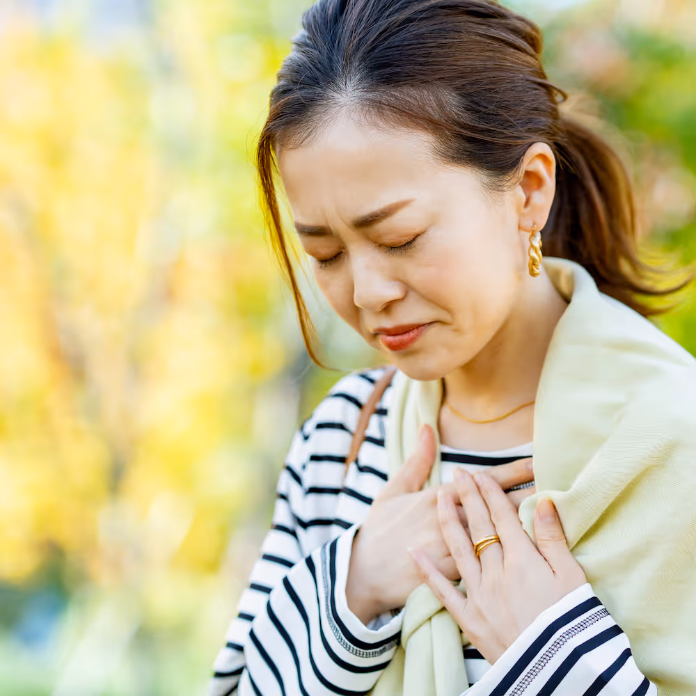 Woman grimacing and holding her chest outdoors against a blurred green and yellow background