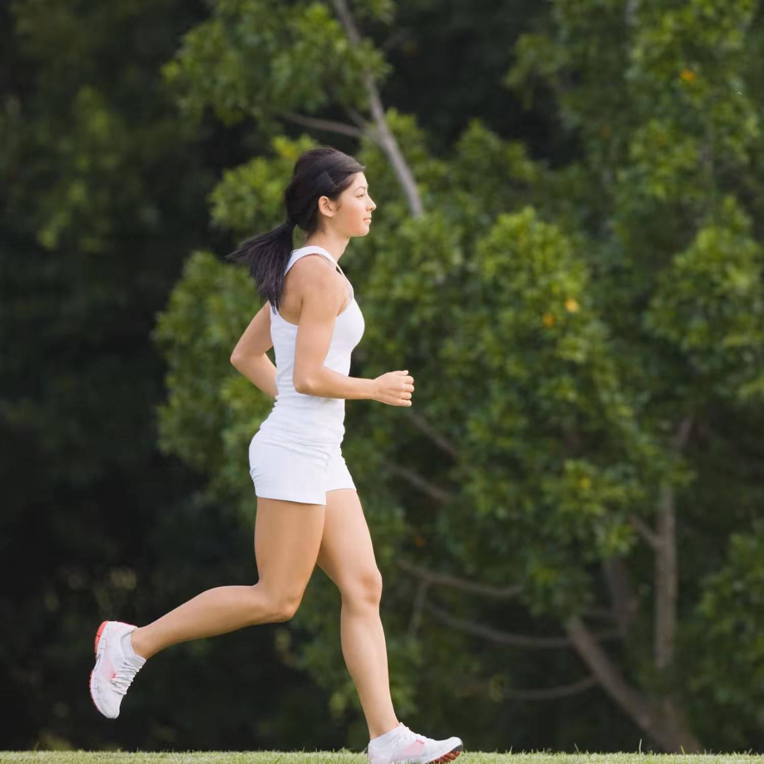 Woman with dark ponytail jogging on grass path in white athletic outfit, surrounded by lush green trees in background