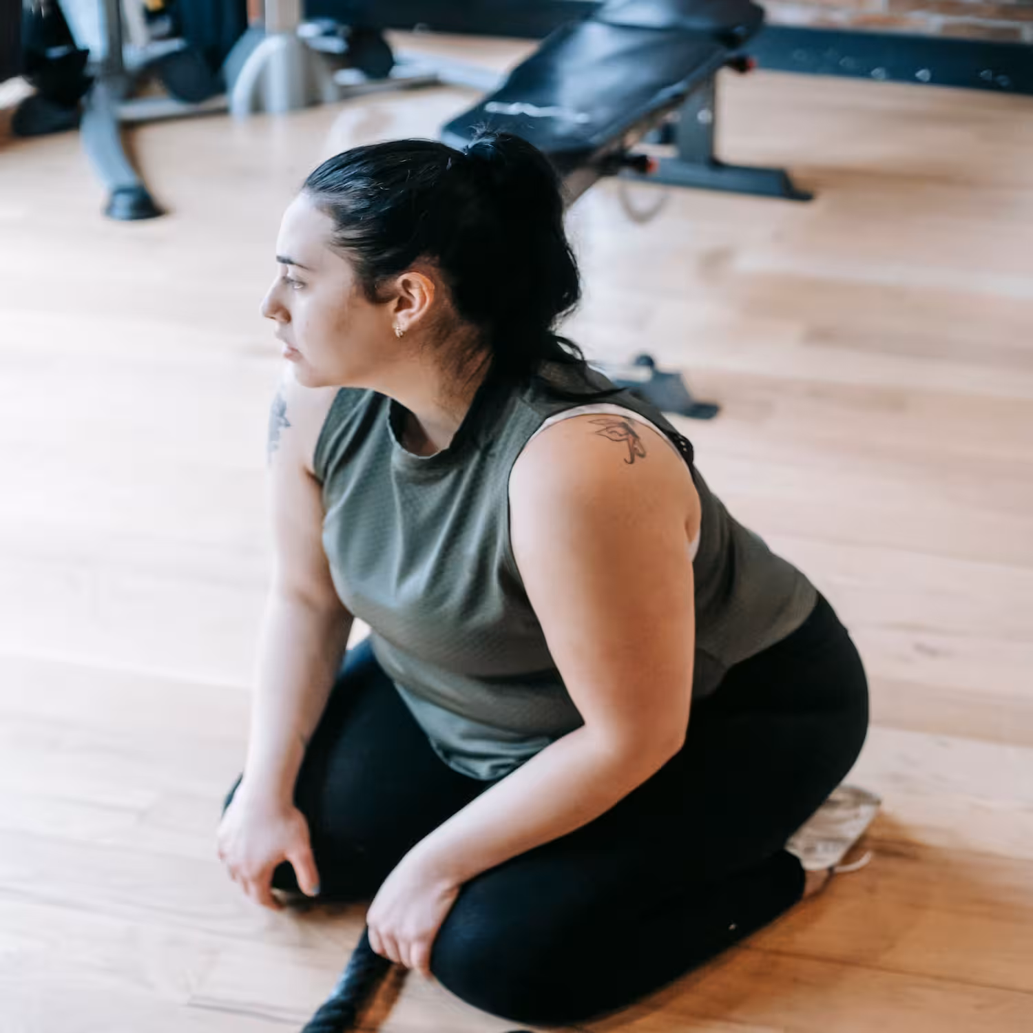 Woman in workout clothes resting on her knees on a gym floor, looking off to the side, with a weight bench behind her