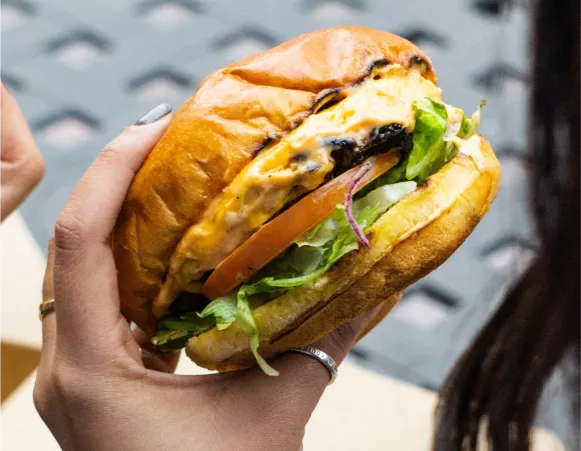 A close-up of a hand holding a cheeseburger with lettuce, tomato, and melted cheese, showcasing the sandwich's layers.
