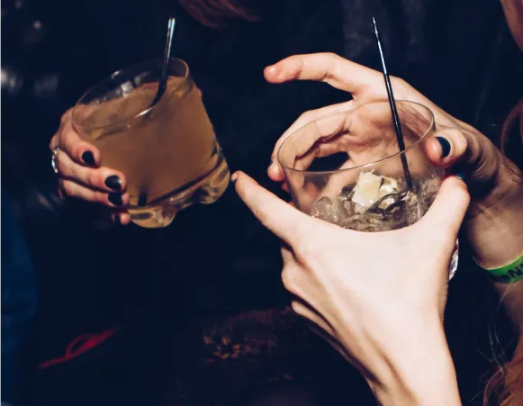 A close-up of two hands holding glasses of cocktails, one with ice and the other with a lighter-colored drink, both featuring straws.