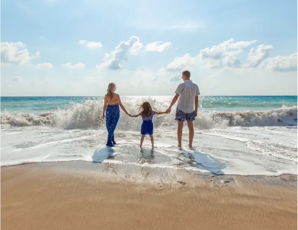 A family of three stands hand in hand at the beach, facing the ocean with waves crashing around them.