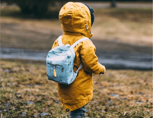 A child wearing a yellow raincoat and a small blue backpack stands outdoors, facing away from the camera.