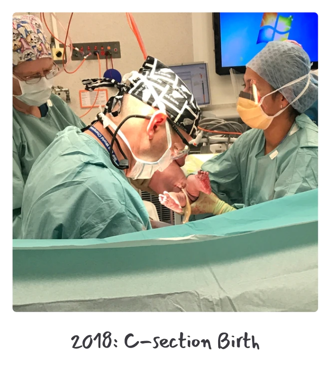 Medical professionals in surgical attire assist in a C-section birth, with one doctor holding a newborn. Monitors and equipment are visible in the background.