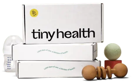 Stack of Tiny Health boxes with a slogan, a baby bottle, and wooden toys.