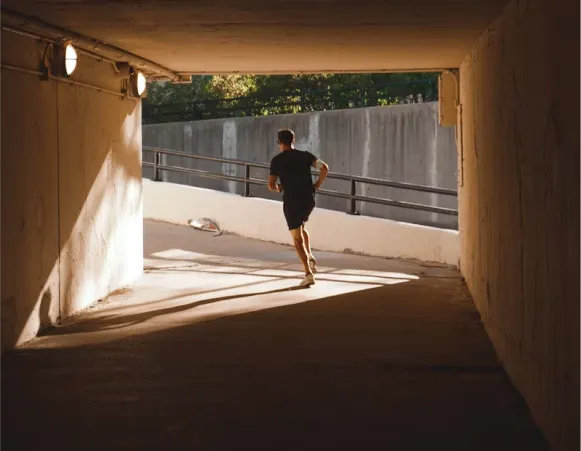 A person running through a tunnel, illuminated by lights, with sunlight streaming in from the opening at the end.