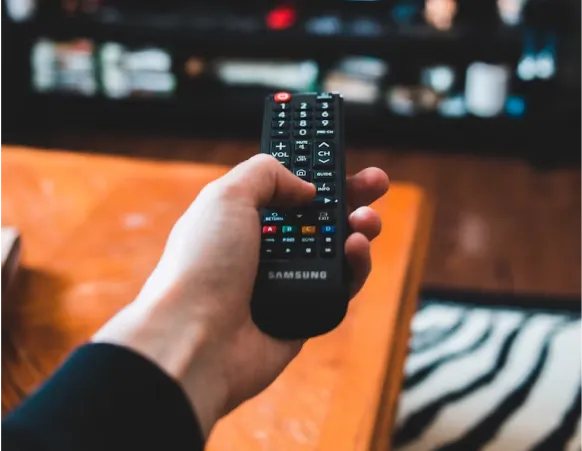 A hand holds a Samsung remote control, with fingers pressing a button, set against a blurred background of a living room.