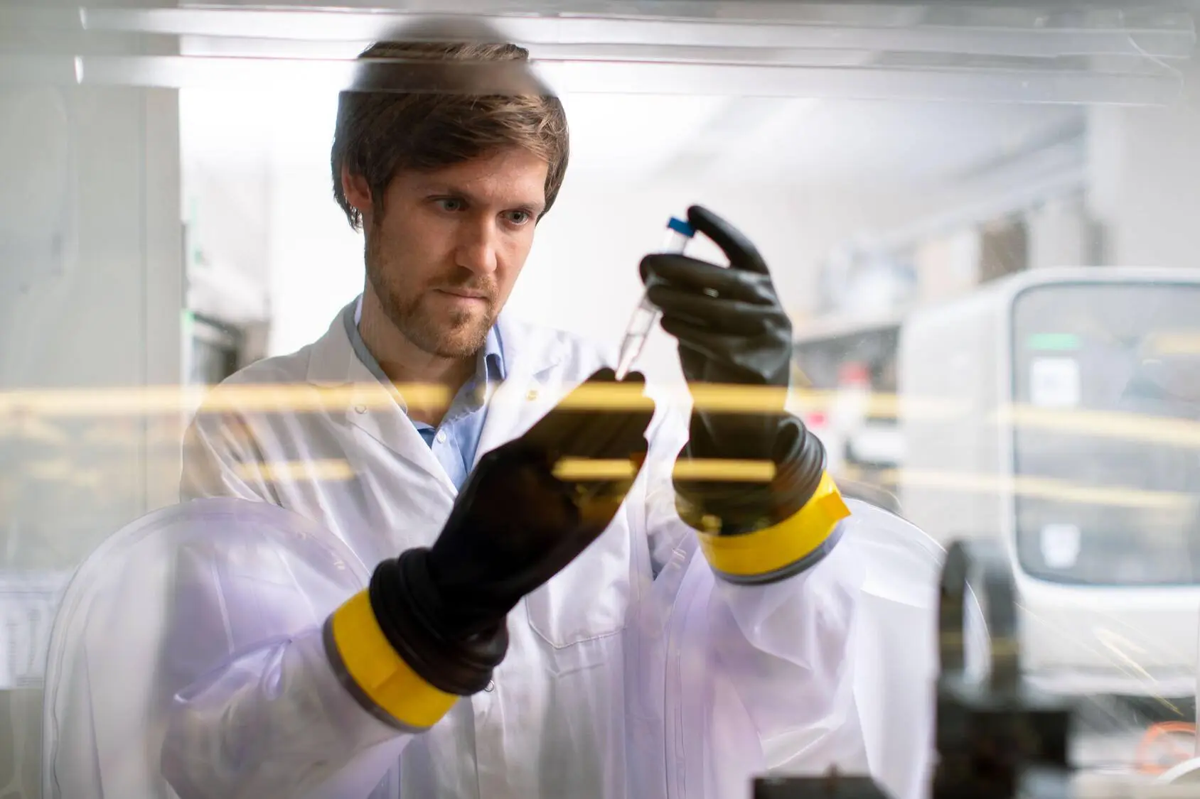 A male scientist in a white lab coat and black gloves examines a test tube in a laboratory setting, emphasizing safety and precision.