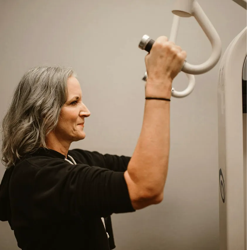 A smiling woman with shoulder-length grey hair exercises by pulling on the handles of a weight machine.