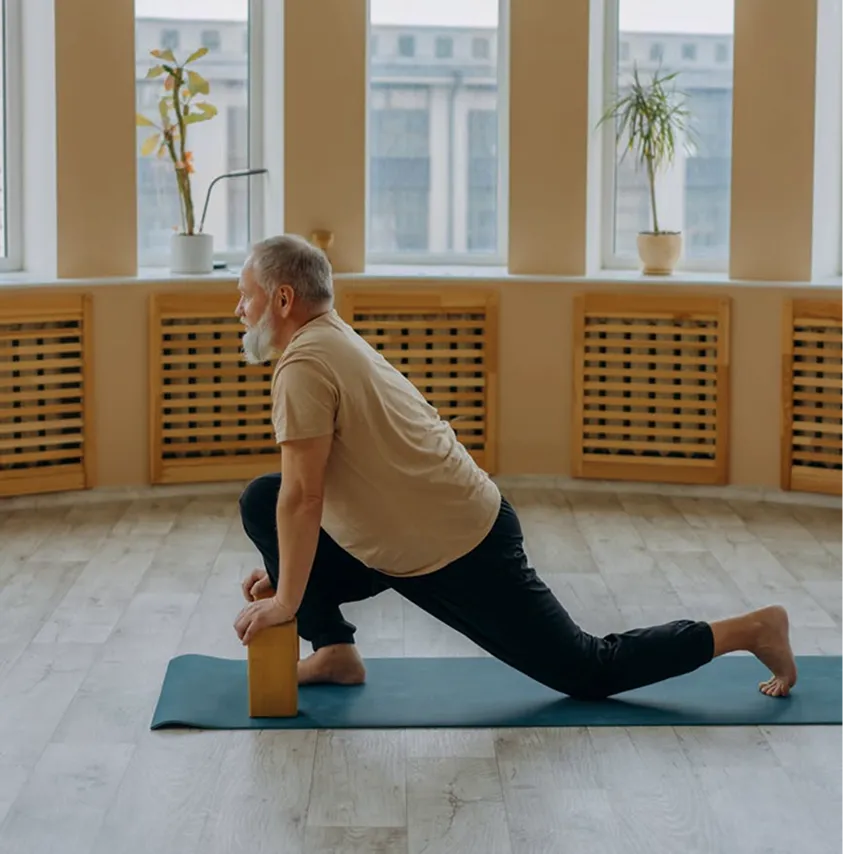 An older man with a grey beard and hair practices a lunge pose on a yoga mat in a sunlit room.
