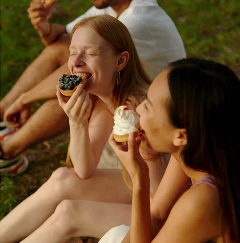Two women sit together on the grass, happily eating small dessert tarts during an outdoor picnic.