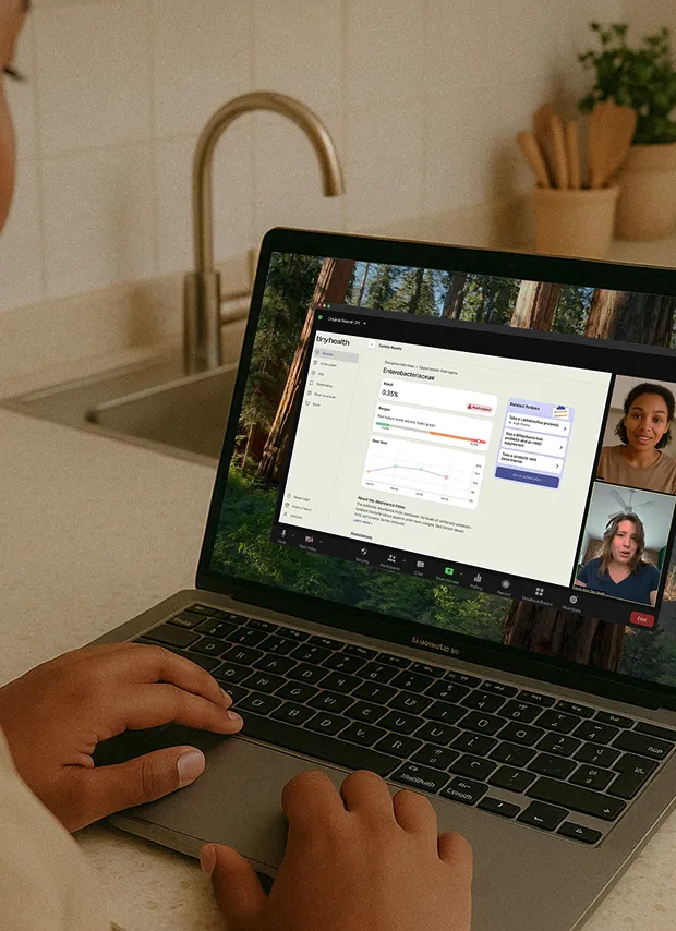 A person at a kitchen counter uses a laptop for a video call. The screen shows a telehealth session where "tinyhealth" results are being reviewed, with two other participants visible in video windows on the side.