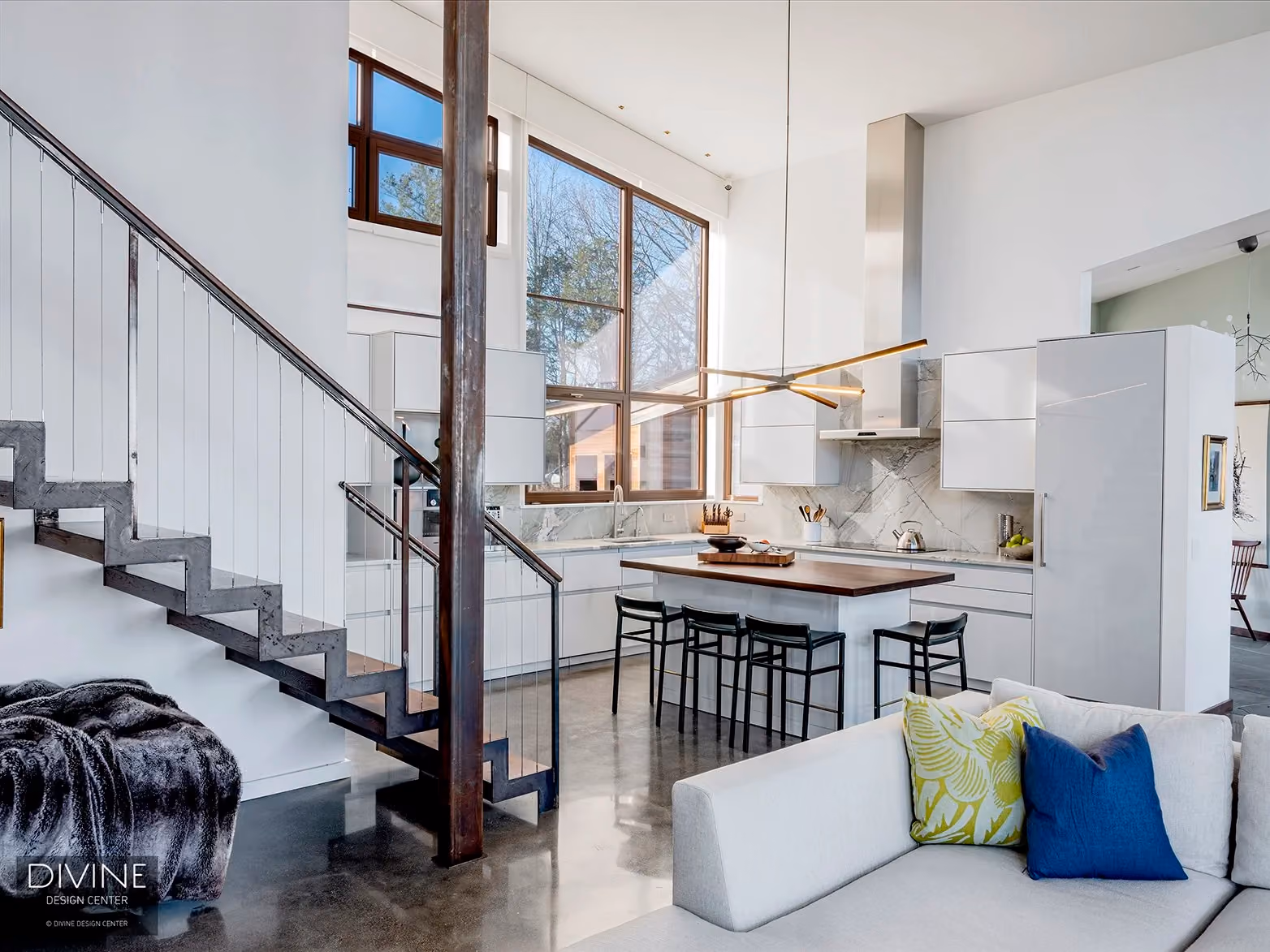 Open kitchen and living area with a black cable-rail staircase, white lacquer cabinetry, a wood dining table with black chairs, and a gray sectional sofa layered with a fringed throw blanket and patterned pillows