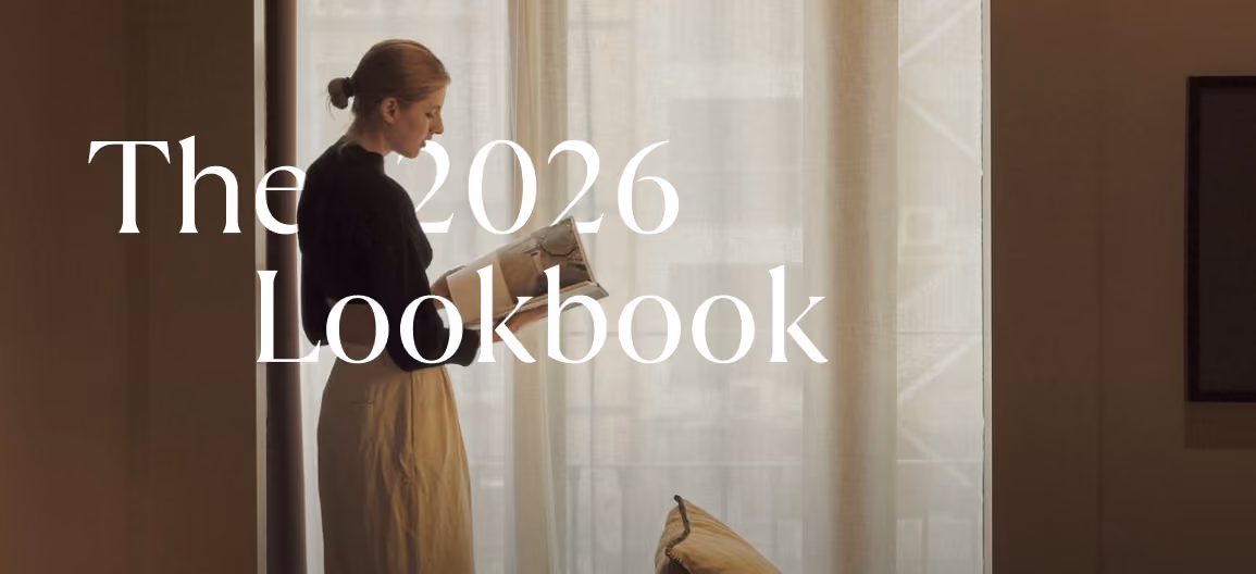Woman in black top and beige skirt reading a book near sheer window curtains.