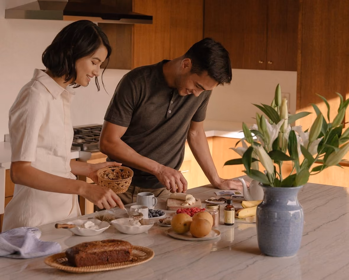 Man and woman preparing food together in a kitchen with ingredients and flowers on the counter.