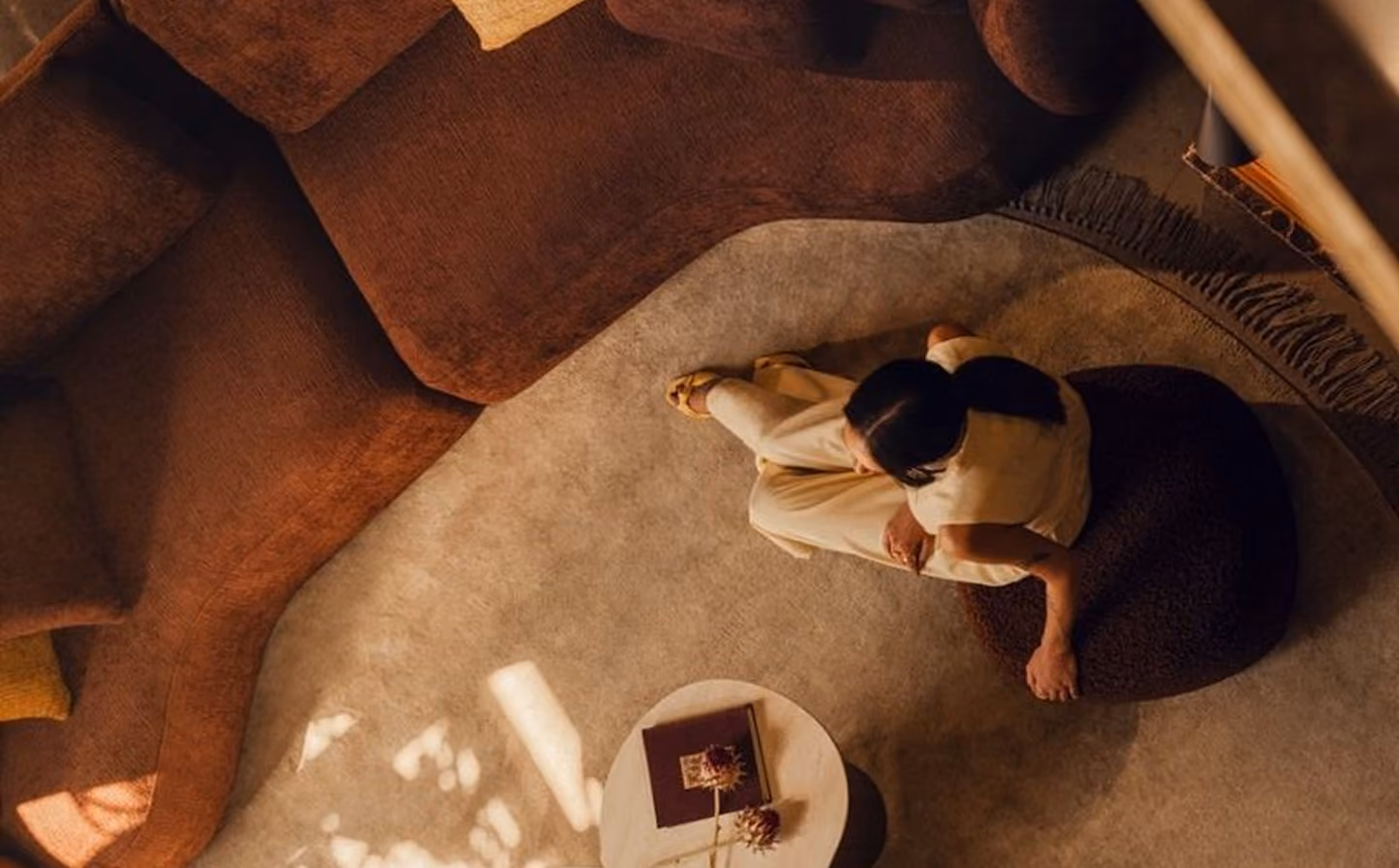 Woman sitting cross-legged on a round cushion on a beige rug beside a brown couch and table.