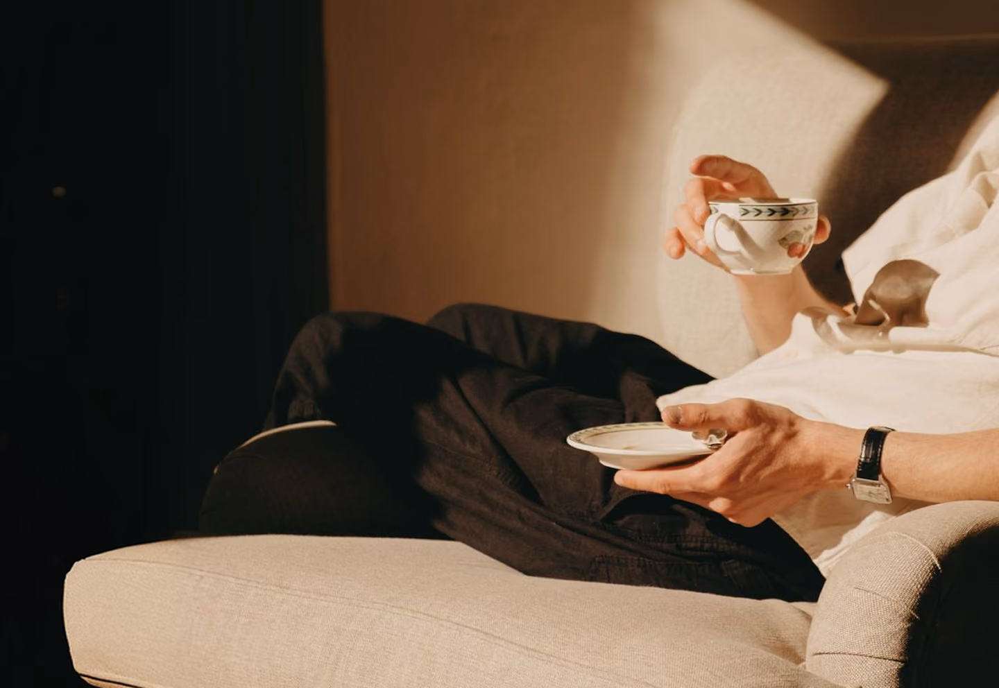 Person sitting on a beige couch holding a teacup and saucer wearing black pants and a white shirt.