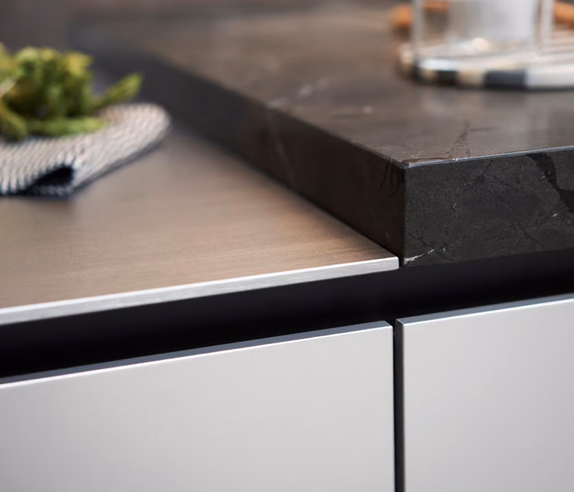 Close-up of a modern kitchen counter showing the edge detail where a black marble countertop overlaps a thinner wood or laminate surface above light gray cabinet doors.