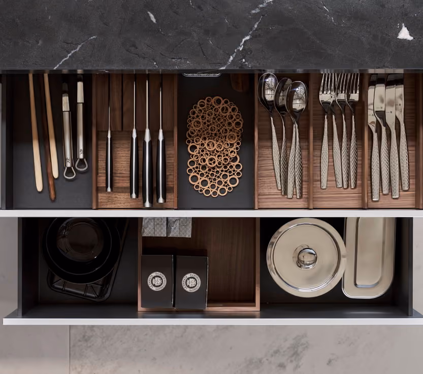 Open kitchen drawers with organized compartments holding chopsticks, bottle openers, kitchen knives, decorative rings, spoons, forks, knives, black plates, two black boxes, and stacked covered metal trays and plates.