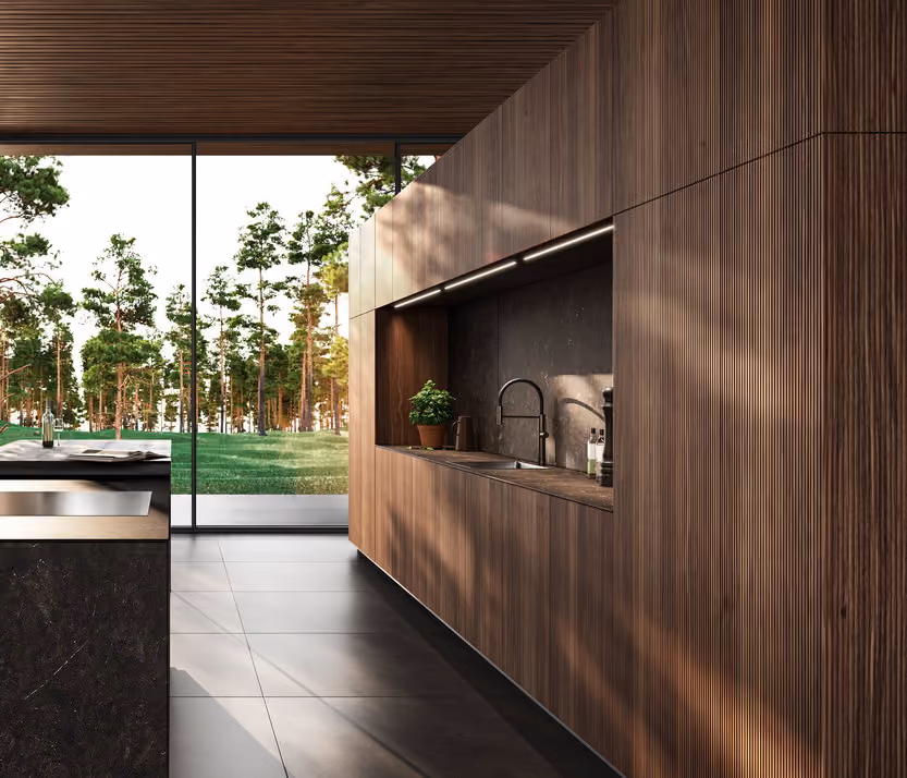 Modern kitchen with dark wood paneled walls and ceiling featuring a recessed sink area, large floor-to-ceiling windows showing a green wooded outdoor landscape, and a dark stone kitchen island with a built-in stovetop.