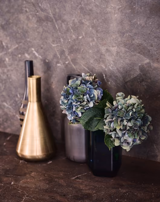 Two blue and green hydrangea flowers with green leaves in a dark rectangular vase on a brown textured surface, with two decorative vases, one metallic gold and one transparent brown, against a gray textured wall background.